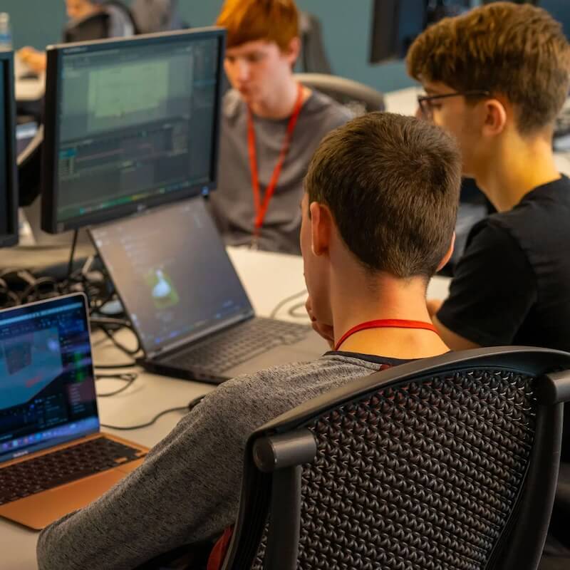 Three young men working on laptops with computer screens displaying coding or digital content in a shared workspace.