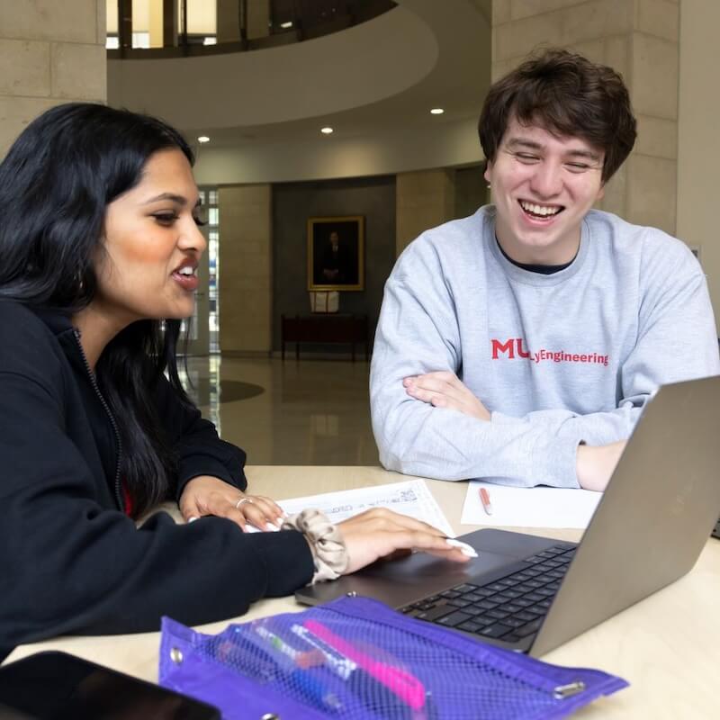 Two students sitting at a table working together on a laptop, with papers and a purple pencil case nearby, inside a building with a modern interior.