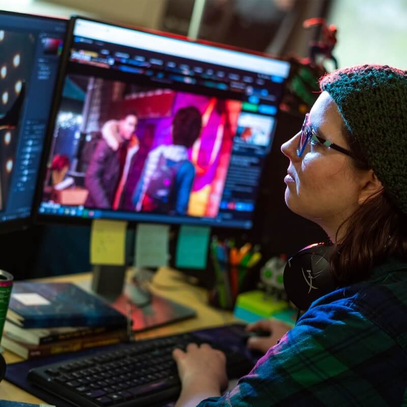 Person wearing a knit cap and glasses working on a computer with dual monitors displaying colorful digital content.