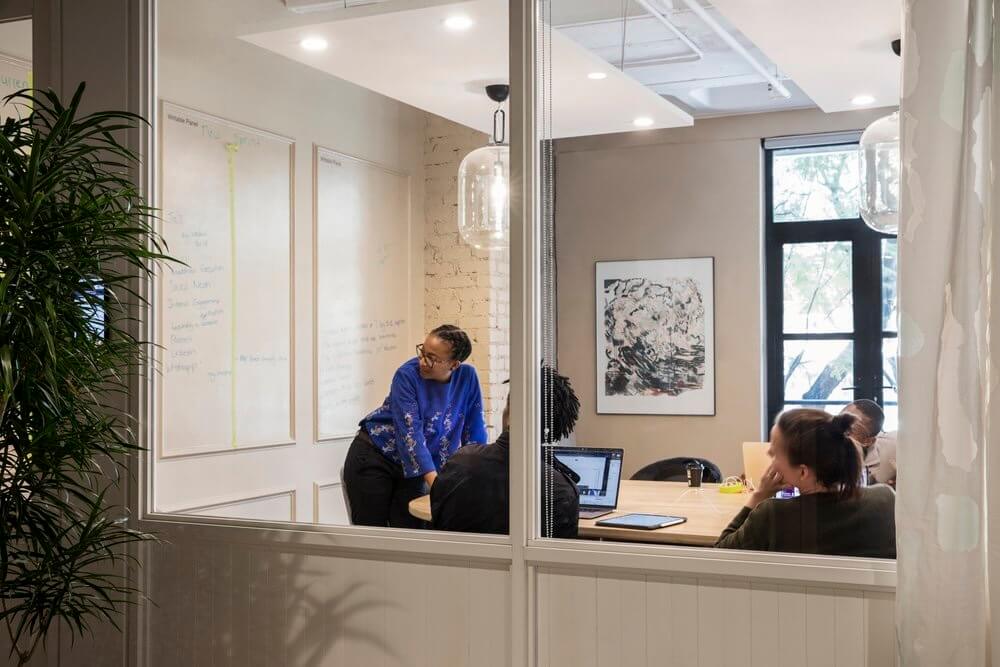 Four people in a modern office meeting room, one standing and leaning on the table while others sit with laptops and tablets.