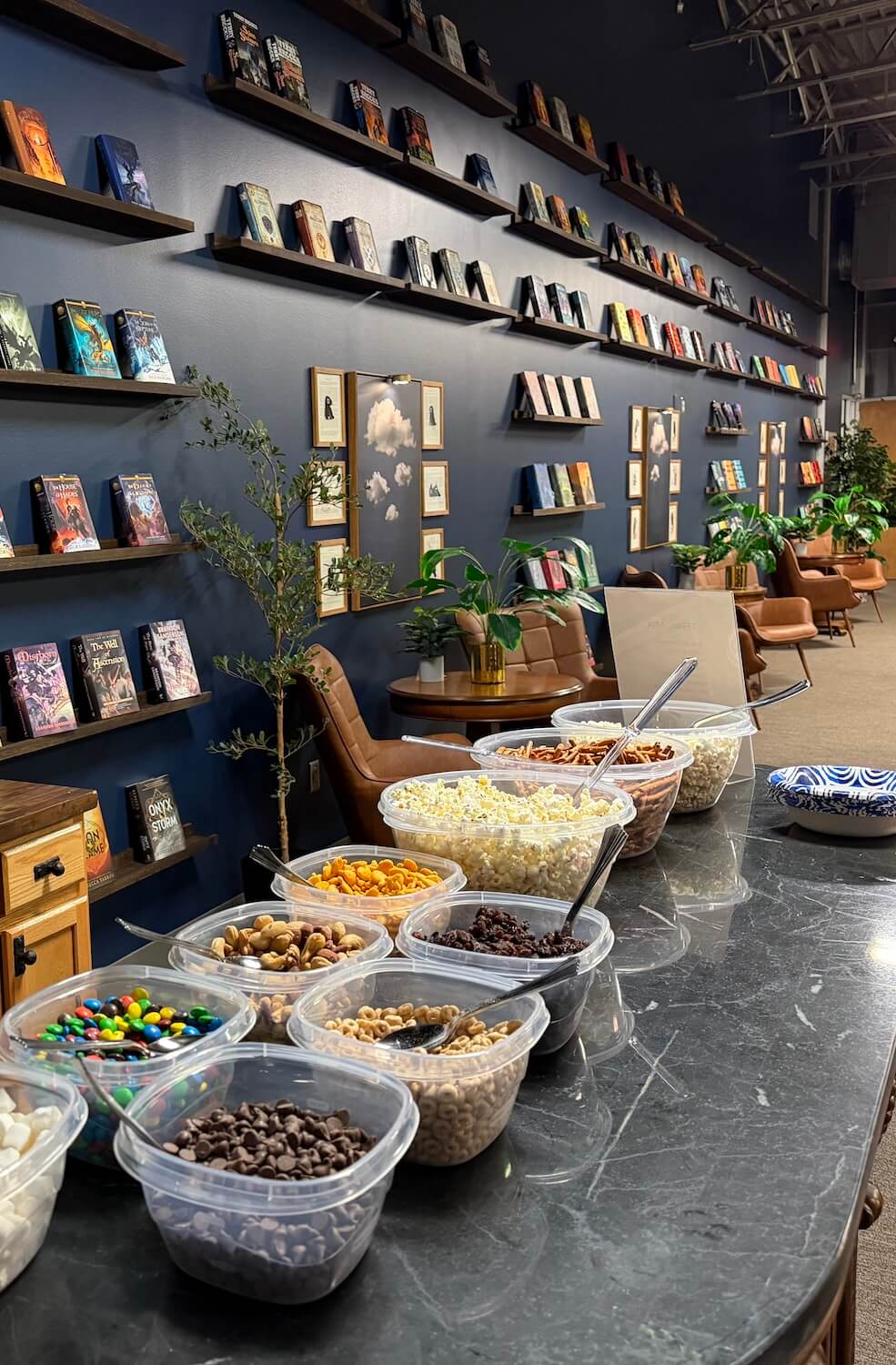 Snack bar with various snacks in plastic containers on a black marble table in a cozy room lined with bookshelves and brown chairs.