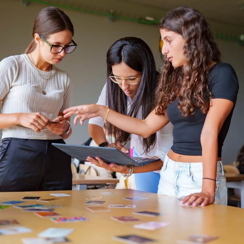 Three young women collaborating over a laptop and phone at a table scattered with cards and papers.