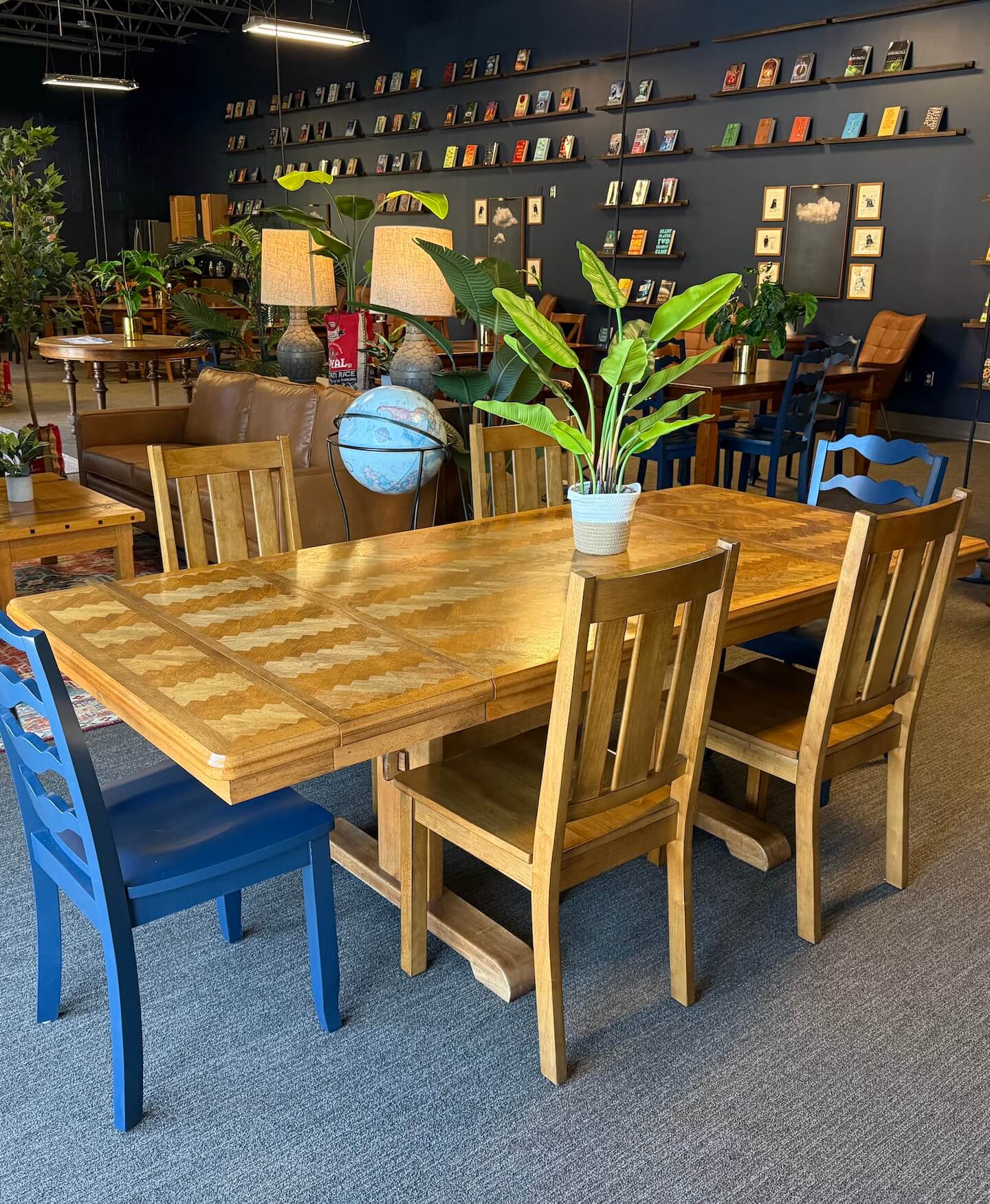 Wooden dining table with a potted plant on top, surrounded by wooden and blue chairs, inside a cozy room with bookshelves and plants.