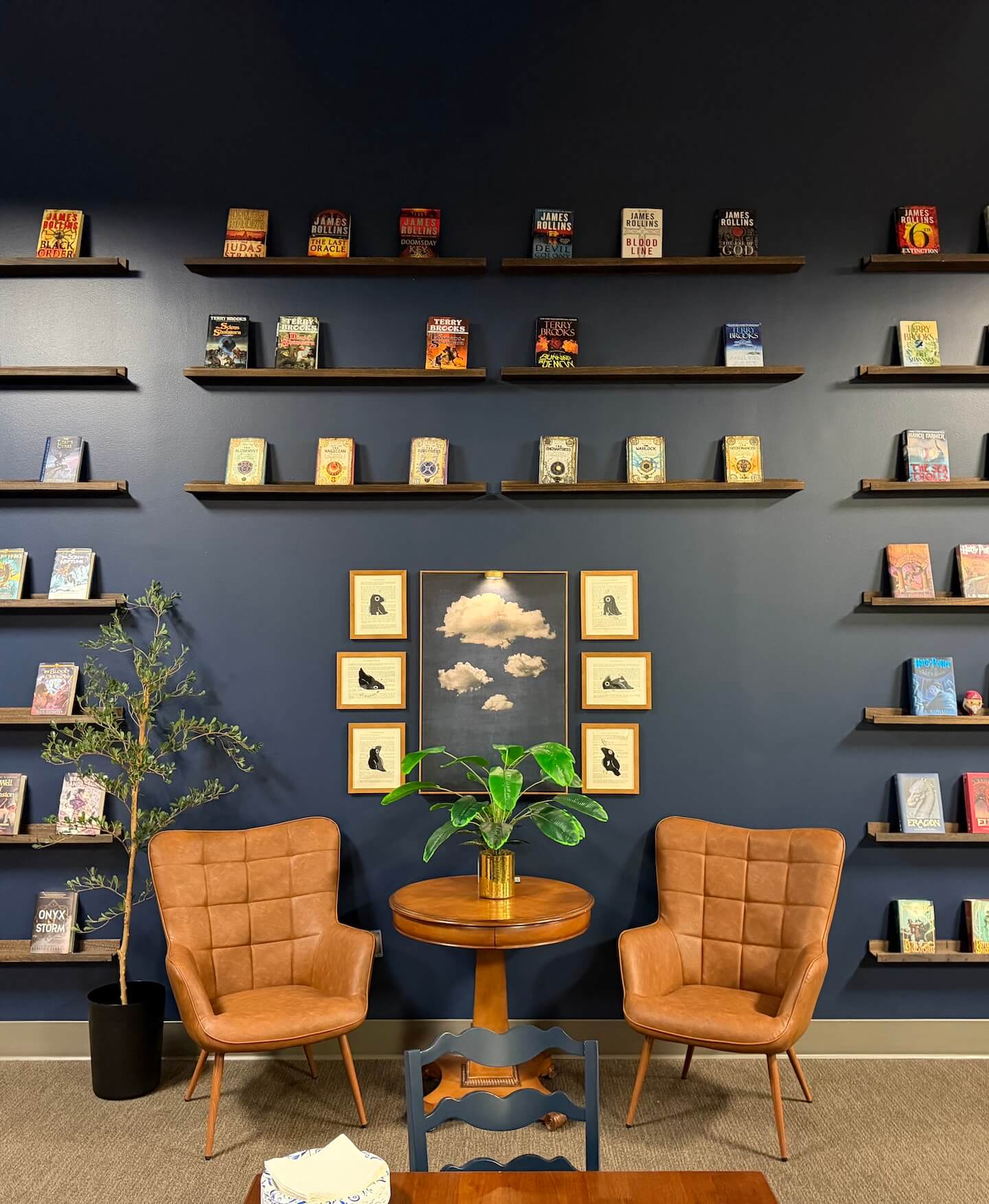 Cozy reading nook with two brown leather chairs, a wooden round table with a green plant, dark blue wall with bookshelves and framed art of clouds and birds.