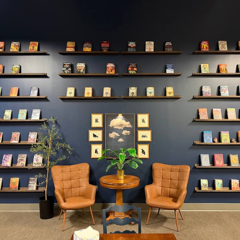 Cozy reading nook with two brown leather armchairs, a round wooden table with a potted plant between them, a tall plant in a black pot, and multiple shelves filled with books on a dark blue wall.