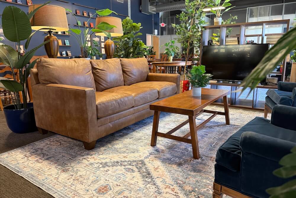 Modern living room with a brown leather sofa, wooden coffee table with a small plant, blue armchairs, a patterned rug, and various green plants and lamps in the background.