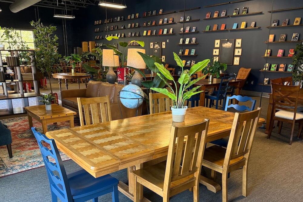 Cozy cafe interior with wooden tables and chairs, plants, a globe, and bookshelves filled with colorful books on a dark blue wall.