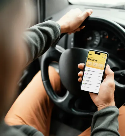 Person holding smartphone showing financial app with account balance while sitting behind car steering wheel.