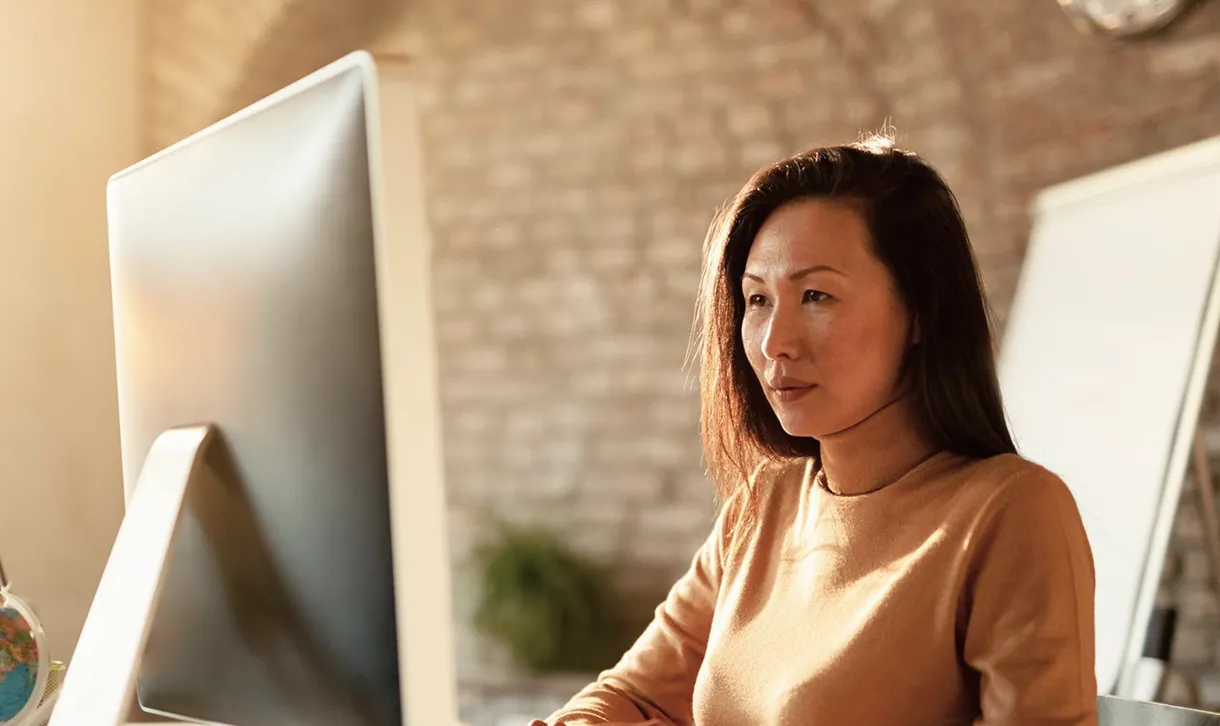Focused woman in a tan sweater working at a desktop computer in a warmly lit office with a brick wall background.