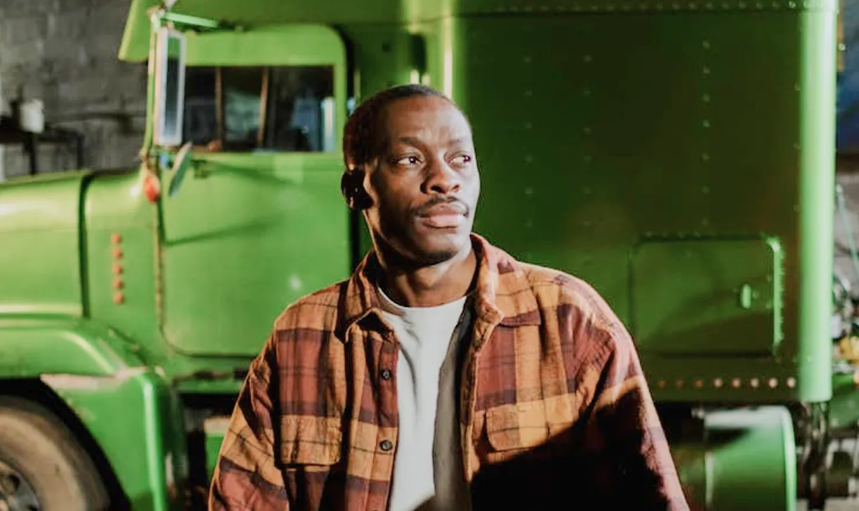 Man wearing a brown and orange checkered shirt standing in front of a green truck inside a garage.