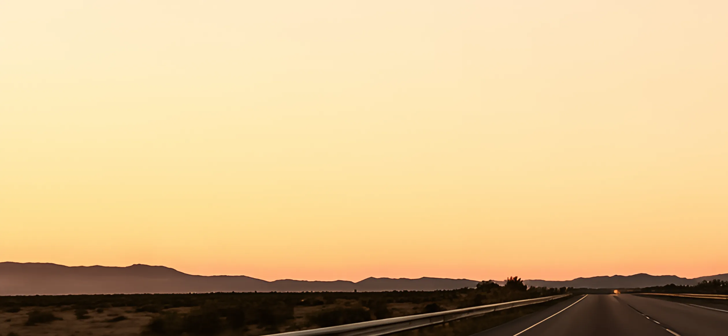 Empty highway with guardrails stretching into the distance under a golden sunset sky with mountains on the horizon.