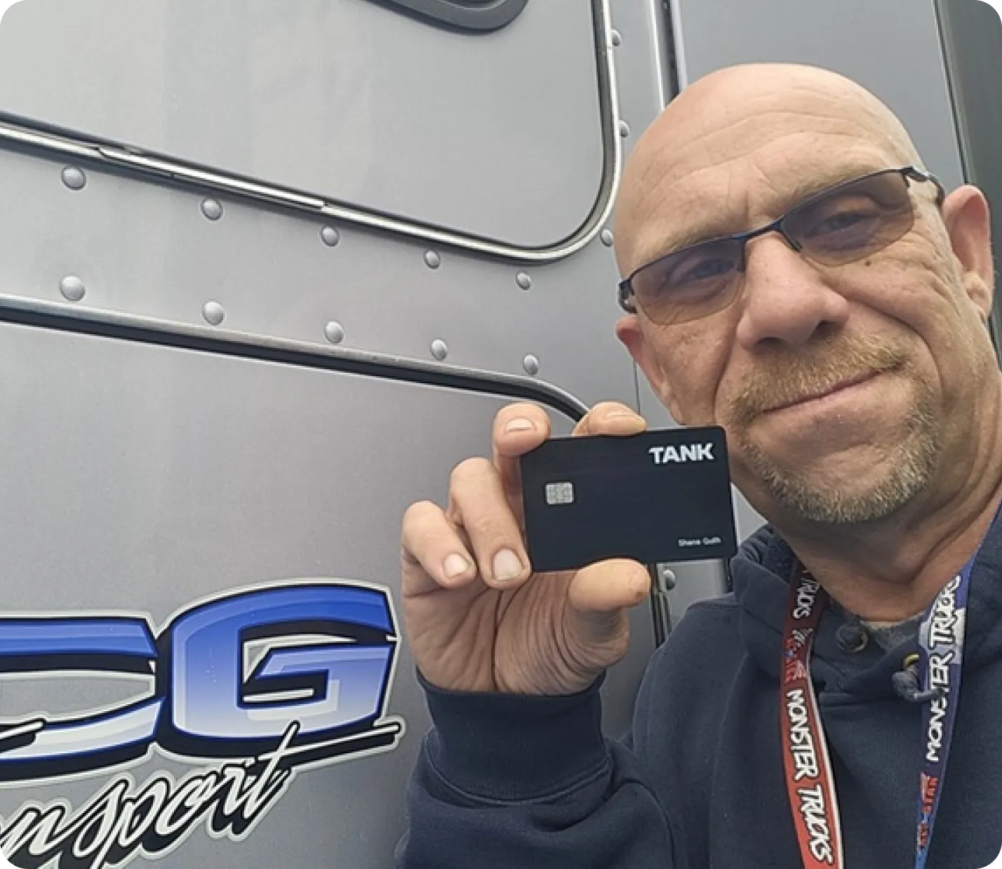 Man wearing glasses holds a black TANK card near a grey truck with blue lettering.