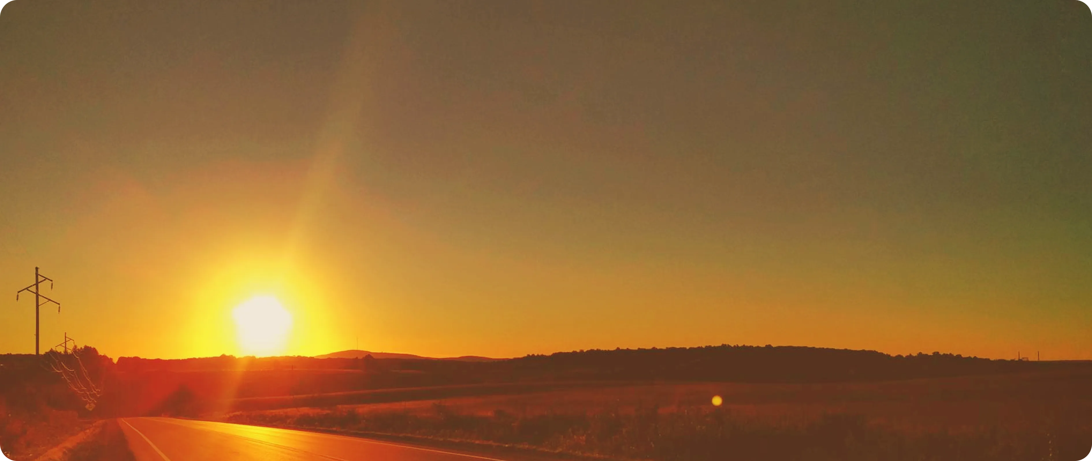 Sun setting over a rural landscape with a road and power lines on the left during golden hour.