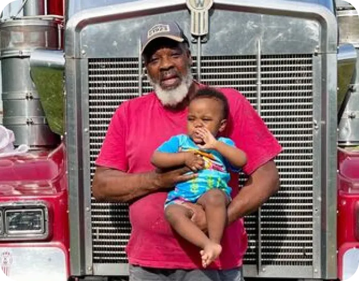 Older man with a gray beard wearing a red shirt and black cap holds a baby in colorful clothes in front of a large red truck.