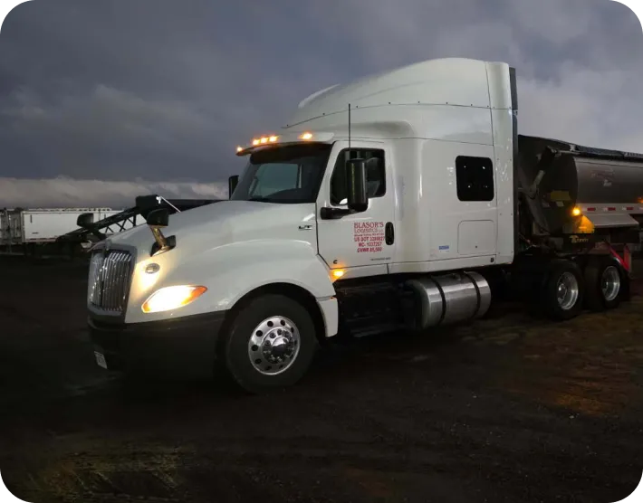 White semi-truck with headlights and roof lights on, parked on muddy ground during dusk or dawn.
