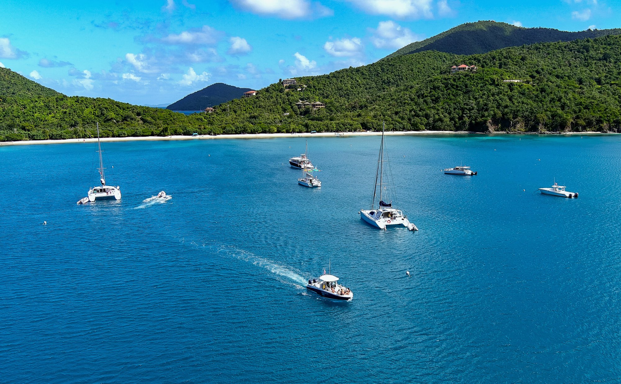 Aerial view of catamarans and boats anchored in turquoise bay with green hillsides and white sand beach USVI