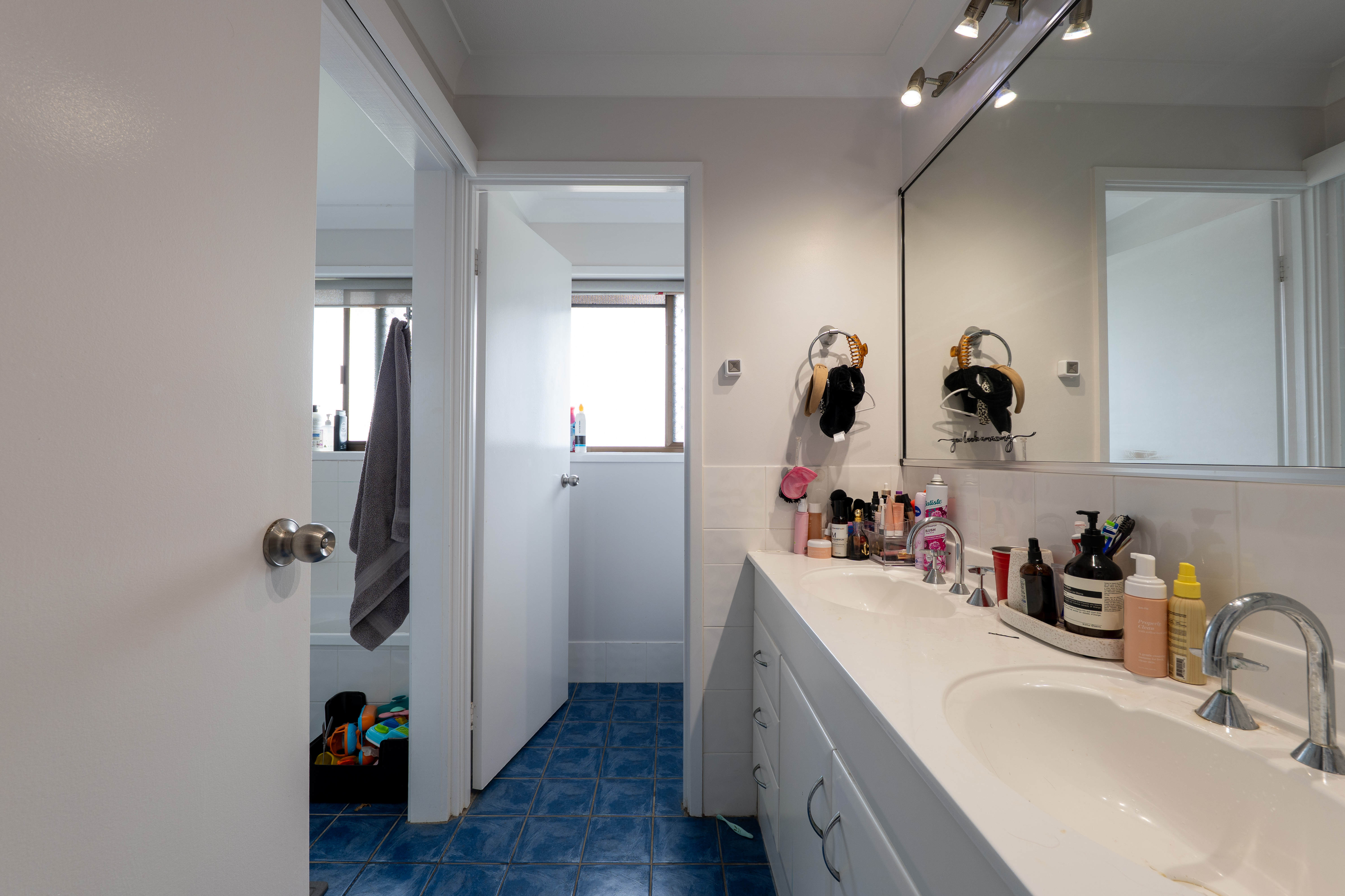 Bathroom with blue tiled floor, white double vanity with assorted toiletries, wall mirror, and an open door leading to another room with toys.