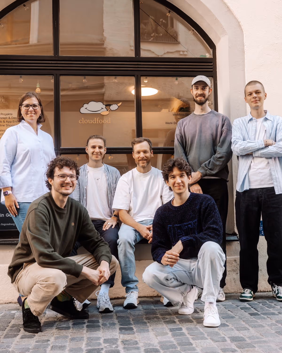 Eine Gruppe von sieben jungen Erwachsenen posiert lächelnd vor einem Fenster mit dem Logo 'cloudfood'.