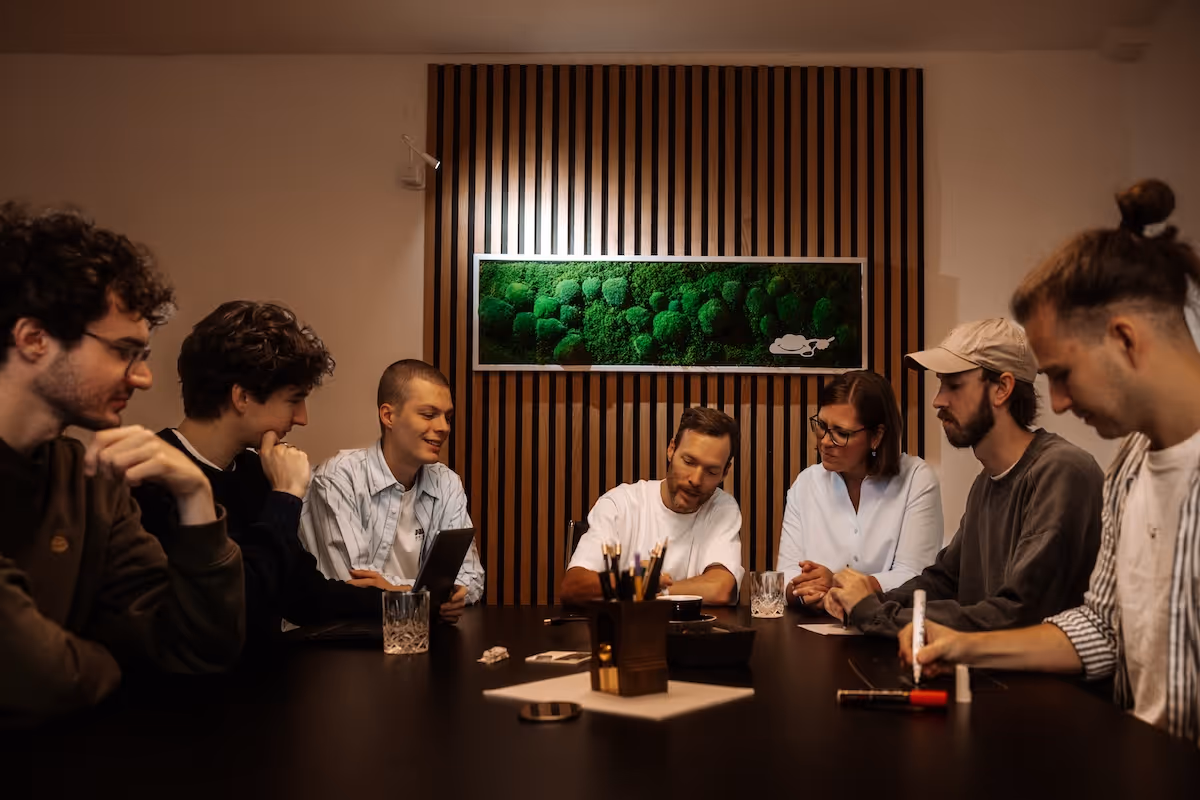 Sieben Menschen sitzen an einem dunklen Tisch und arbeiten gemeinsam in einem modernen Raum mit Holzlamellen und einem Wandbild aus Moos.