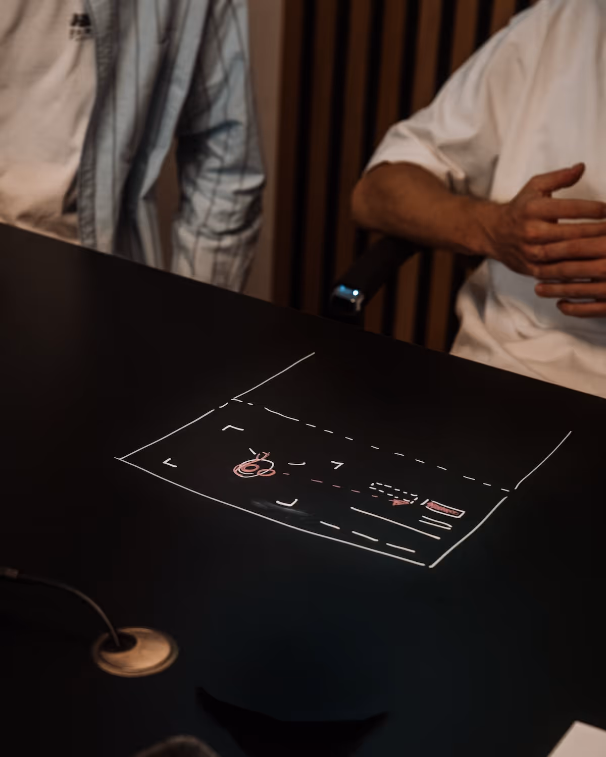 Zwei Personen, die vor einem schwarzen Tisch sitzen, auf dem ein strategisches Diagramm mit Pfeilen und Kreisen gezeichnet ist.