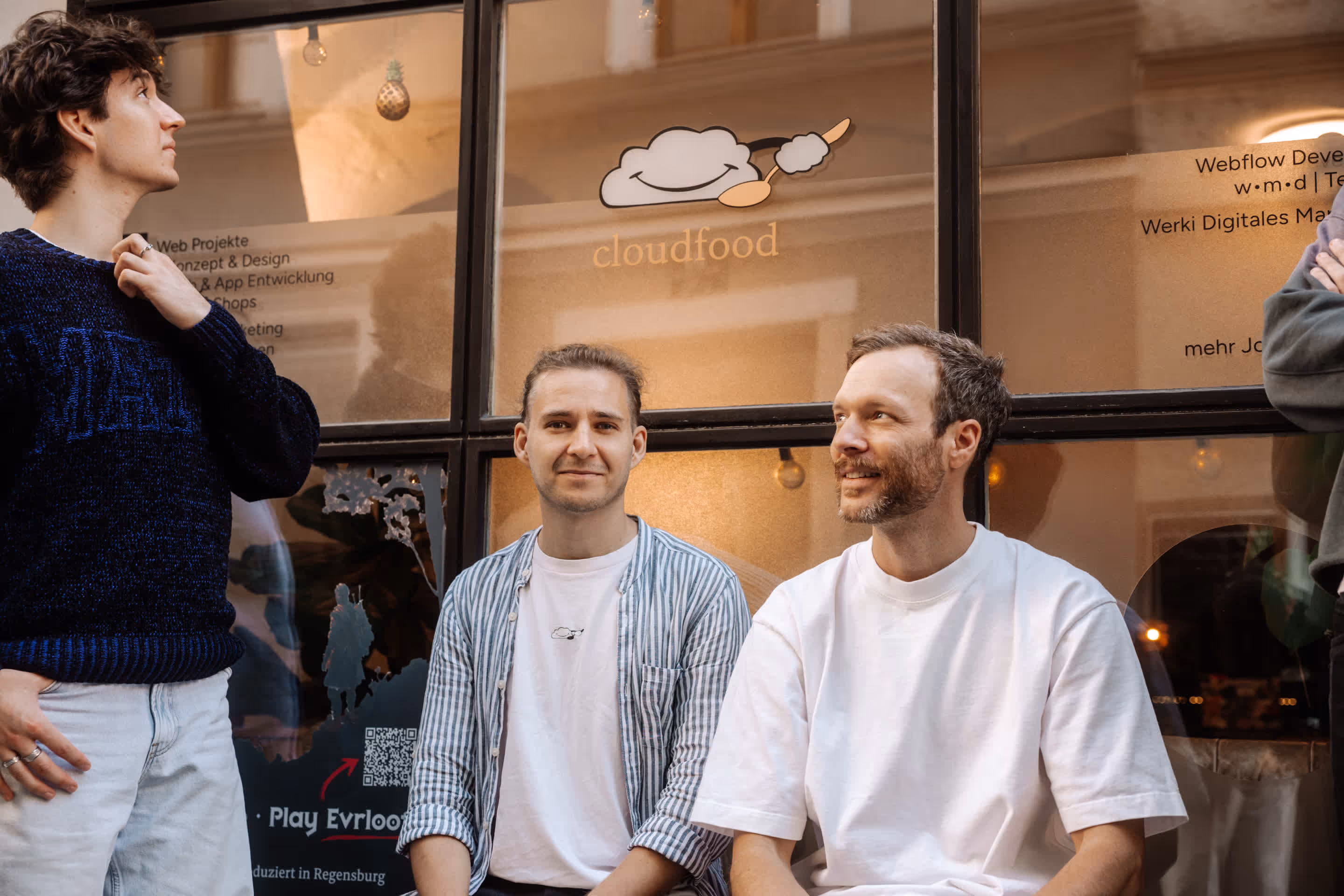 Drei Männer vor einem Fenster mit dem Logo 'cloudfood', zwei sitzen und einer steht am linken Bildrand.
