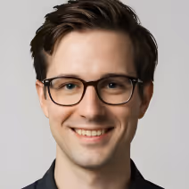 Smiling young man with dark hair and glasses wearing a dark shirt against a light gray background.
