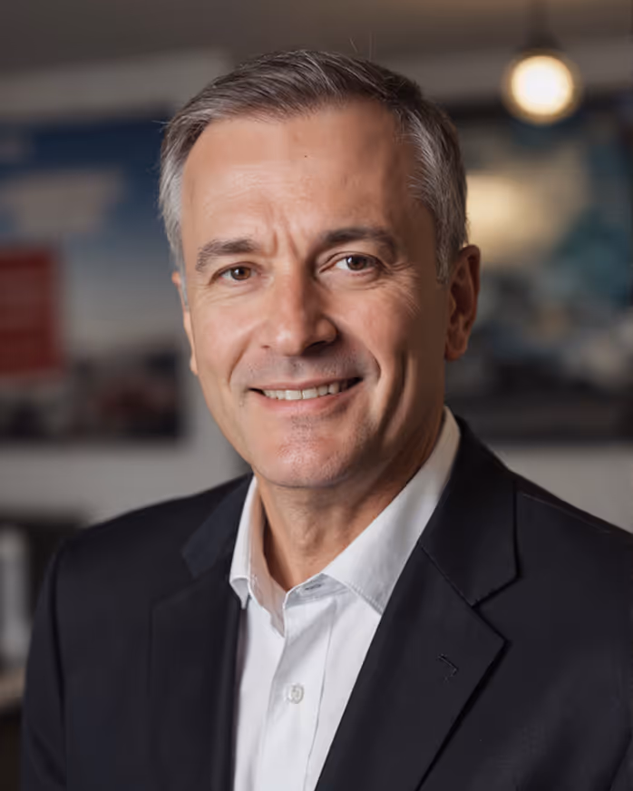 Smiling middle-aged man with gray hair wearing a black blazer and white shirt in an indoor setting.