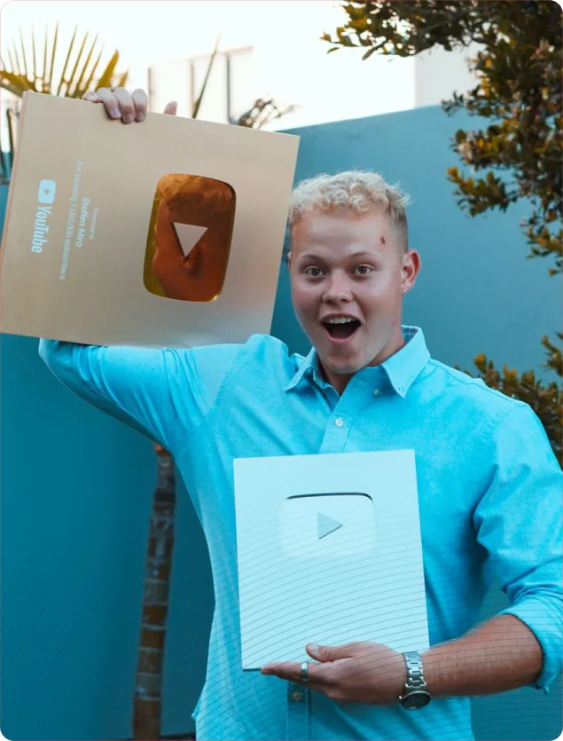 Smiling young man holding a gold and a silver YouTube Play Button award outdoors.