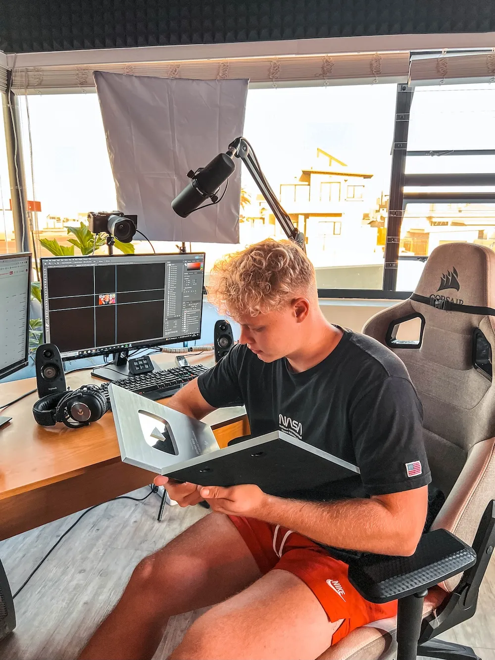 Young man sitting in a gaming chair holding and looking at a silver YouTube Play Button award in a bright room with computer monitors and podcasting equipment.