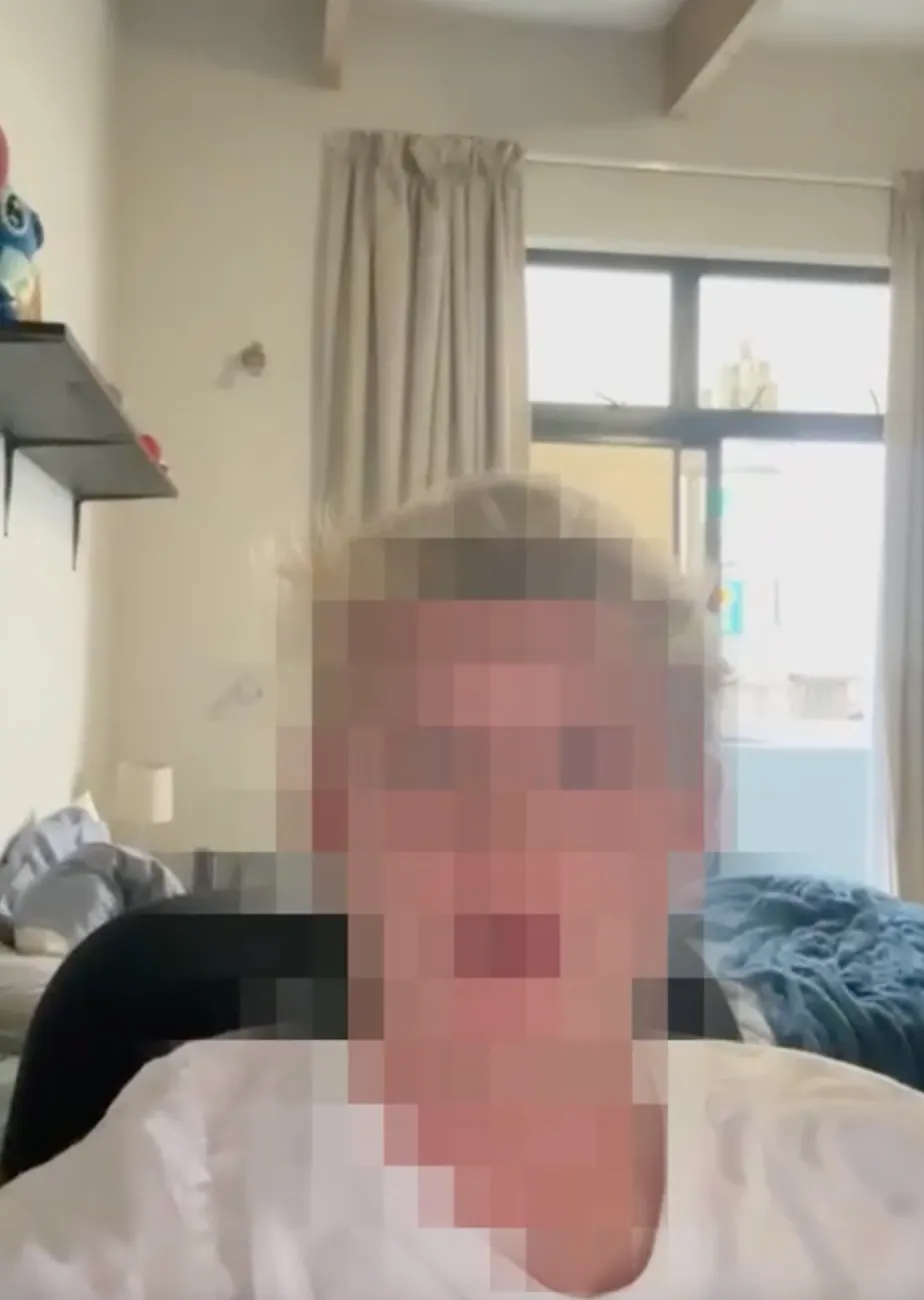 Blurred person sitting indoors with a shelf and window in the background.
