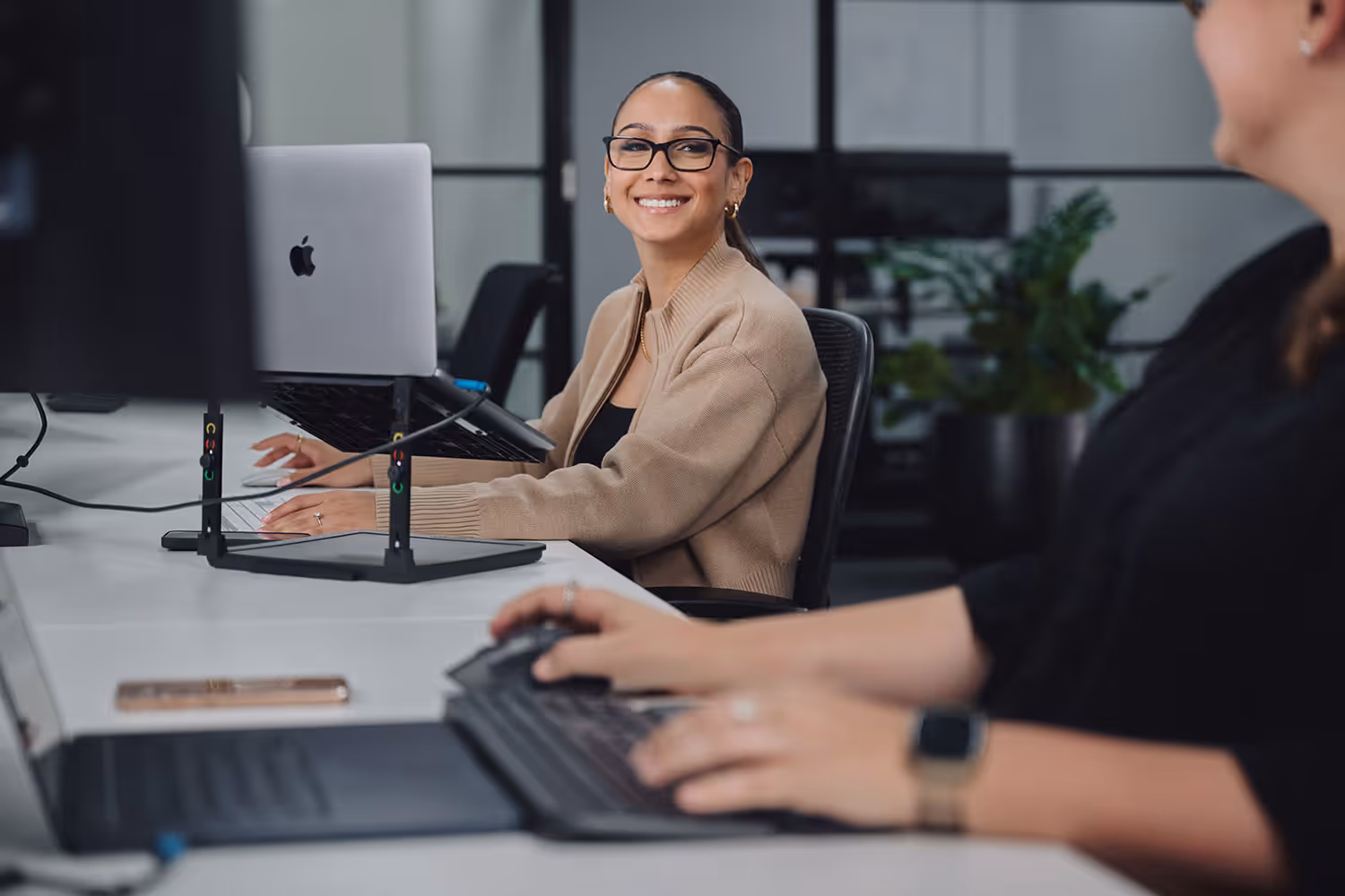 Smiling woman with glasses and beige cardigan sitting at a desk with a laptop in a modern office.