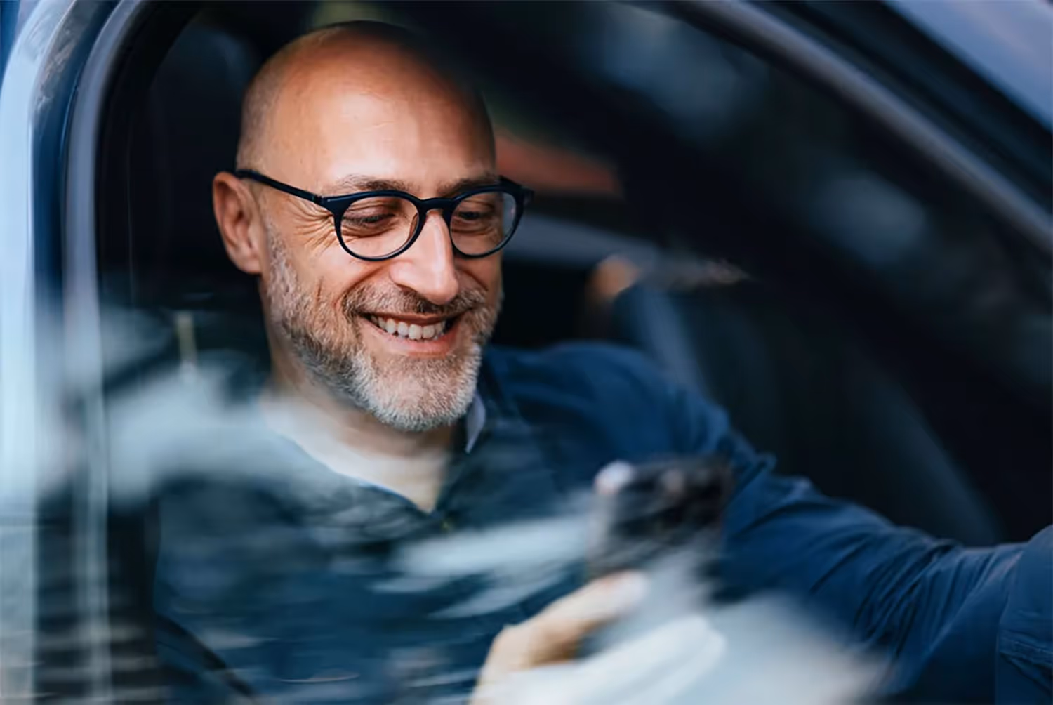 Smiling bald man with glasses sitting in a car looking at his phone.