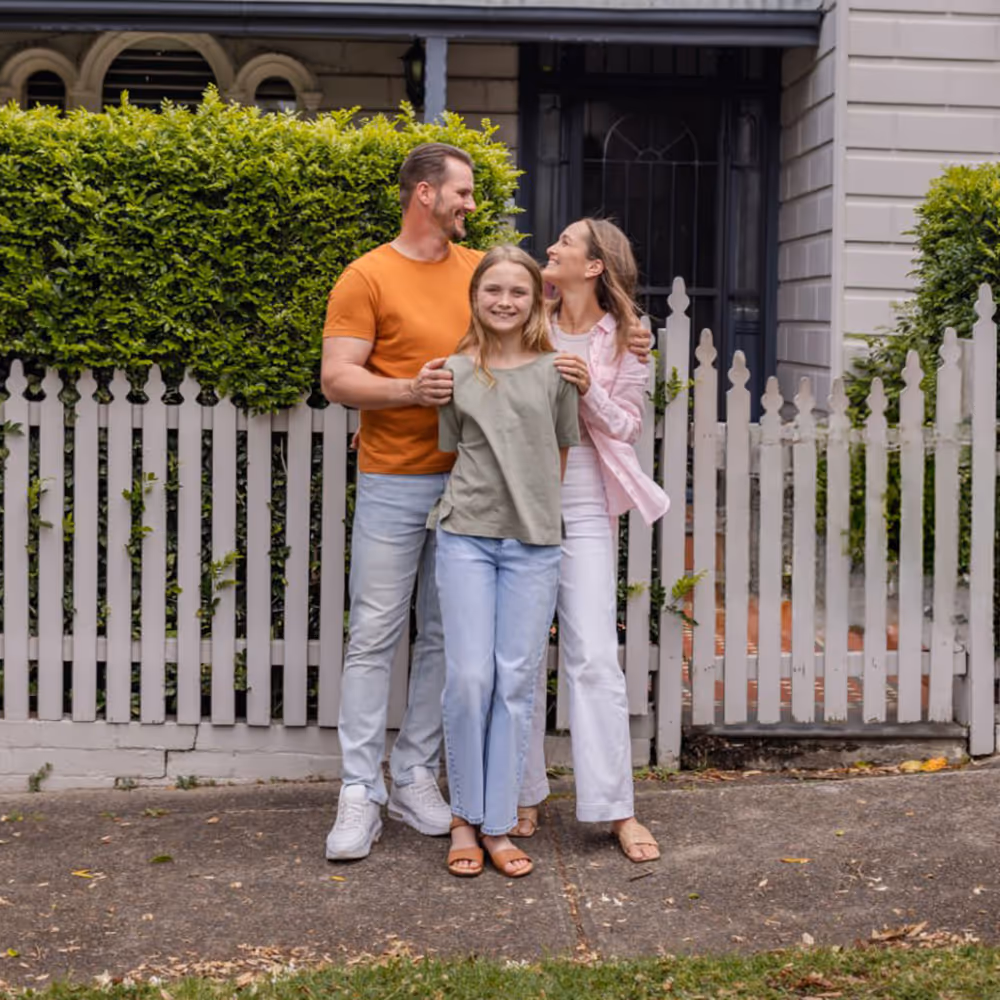 Smiling family of two adults and a child standing together on a sidewalk in front of a white picket fence and green bushes.