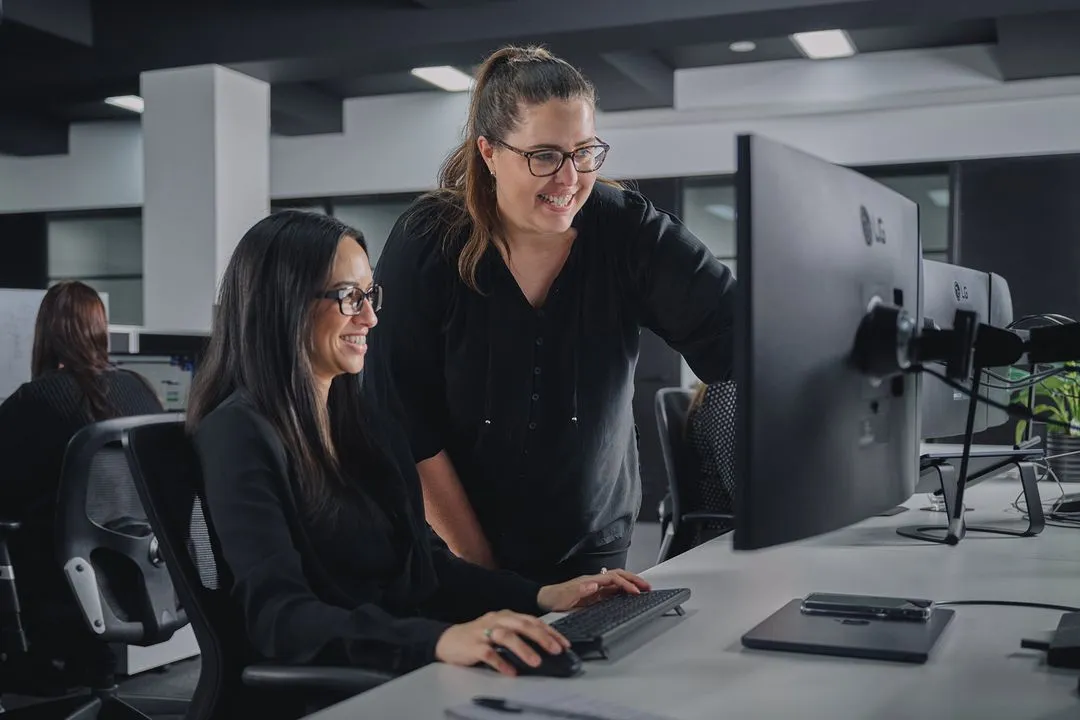 Two colleagues smiling and collaborating at a computer in an open office environment.