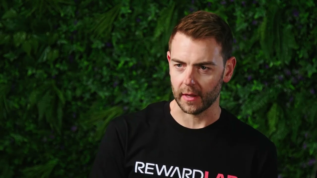 Man with short brown hair and beard wearing a black RewardLab T-shirt standing against a green leafy background.