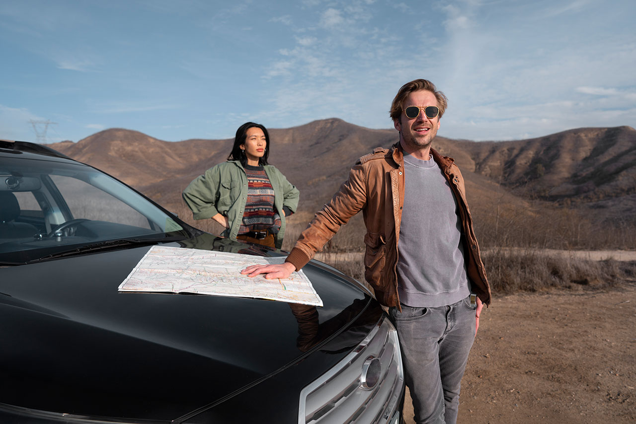 Man and woman standing beside a black car outdoors with a map on the car hood and mountains in the background.