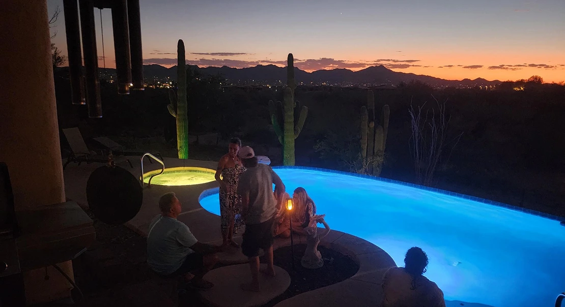 Small group of people gathered beside a pool at sunset with desert mountains in the background.