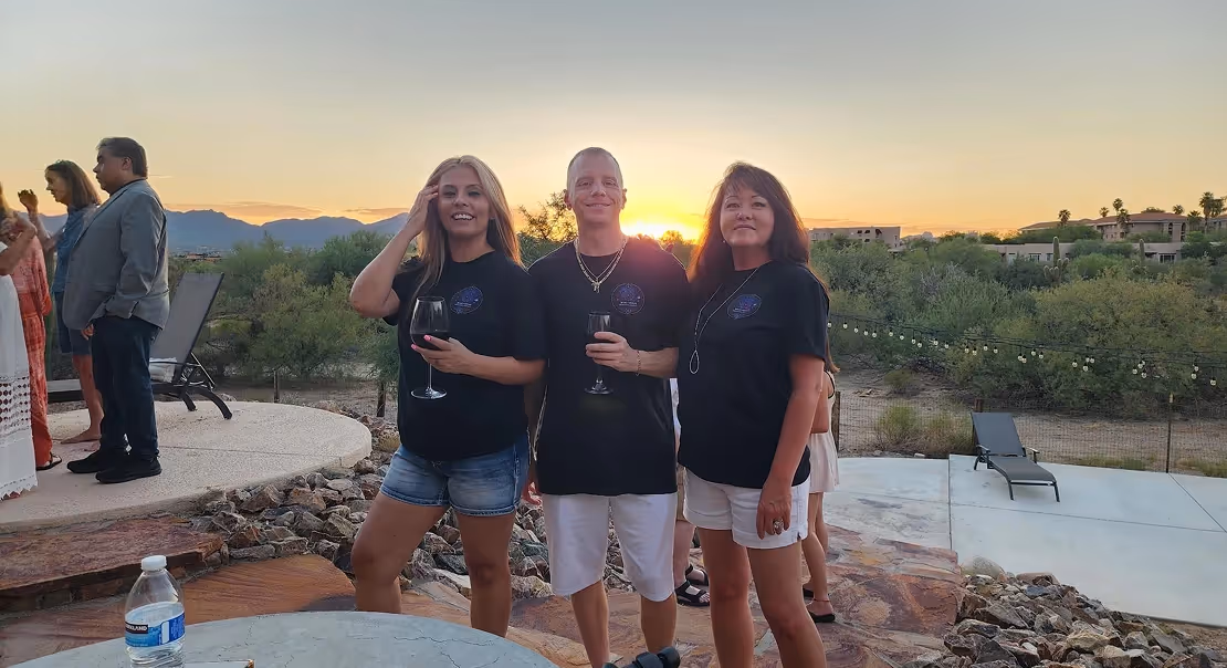 Three people standing outdoors at sunset, posing with wine glasses.