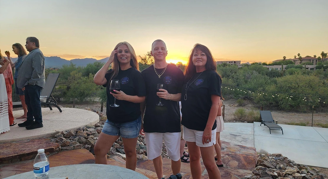 Three people standing outdoors at sunset, posing with wine glasses.
