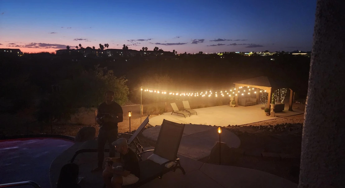 Evening view of a backyard patio with lounge chairs, string lights, and a gazebo illuminated against the night sky.