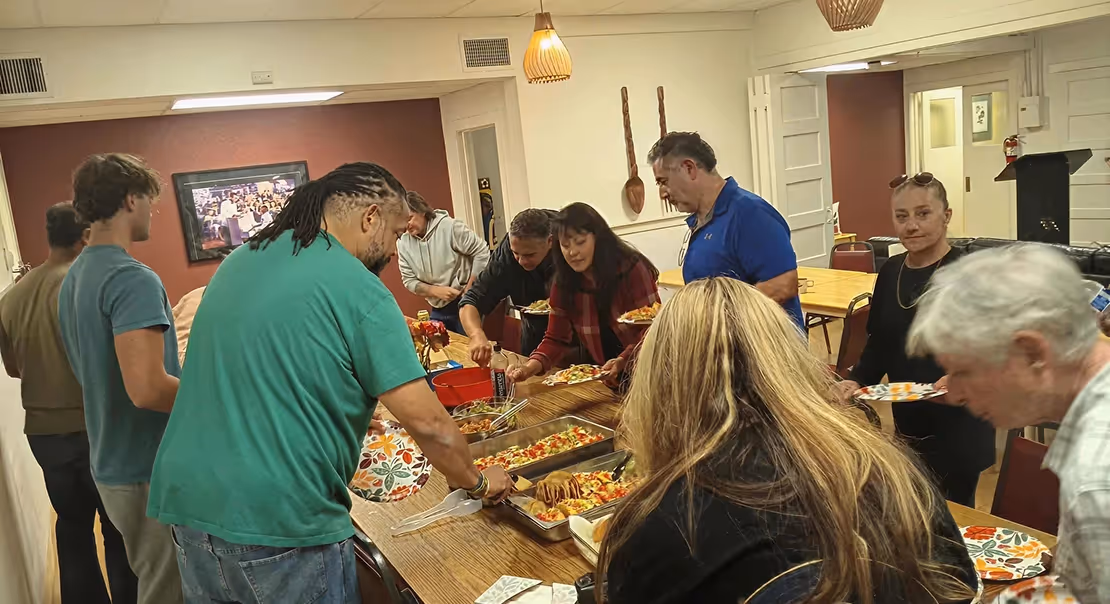 People gathered around a long table indoors, serving themselves food from shared dishes during a meal.