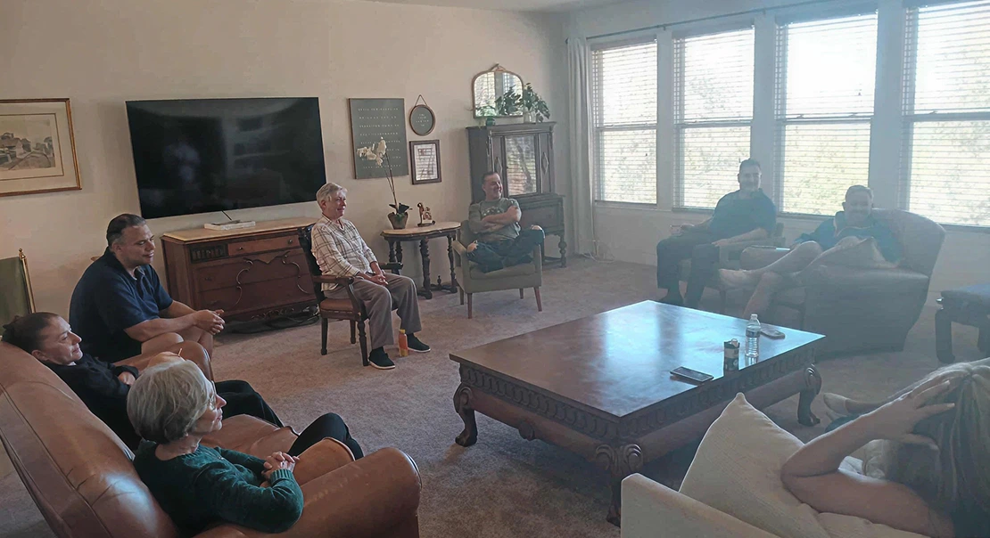 Several adults sitting in a living room arranged in a circle, engaged in a group discussion.