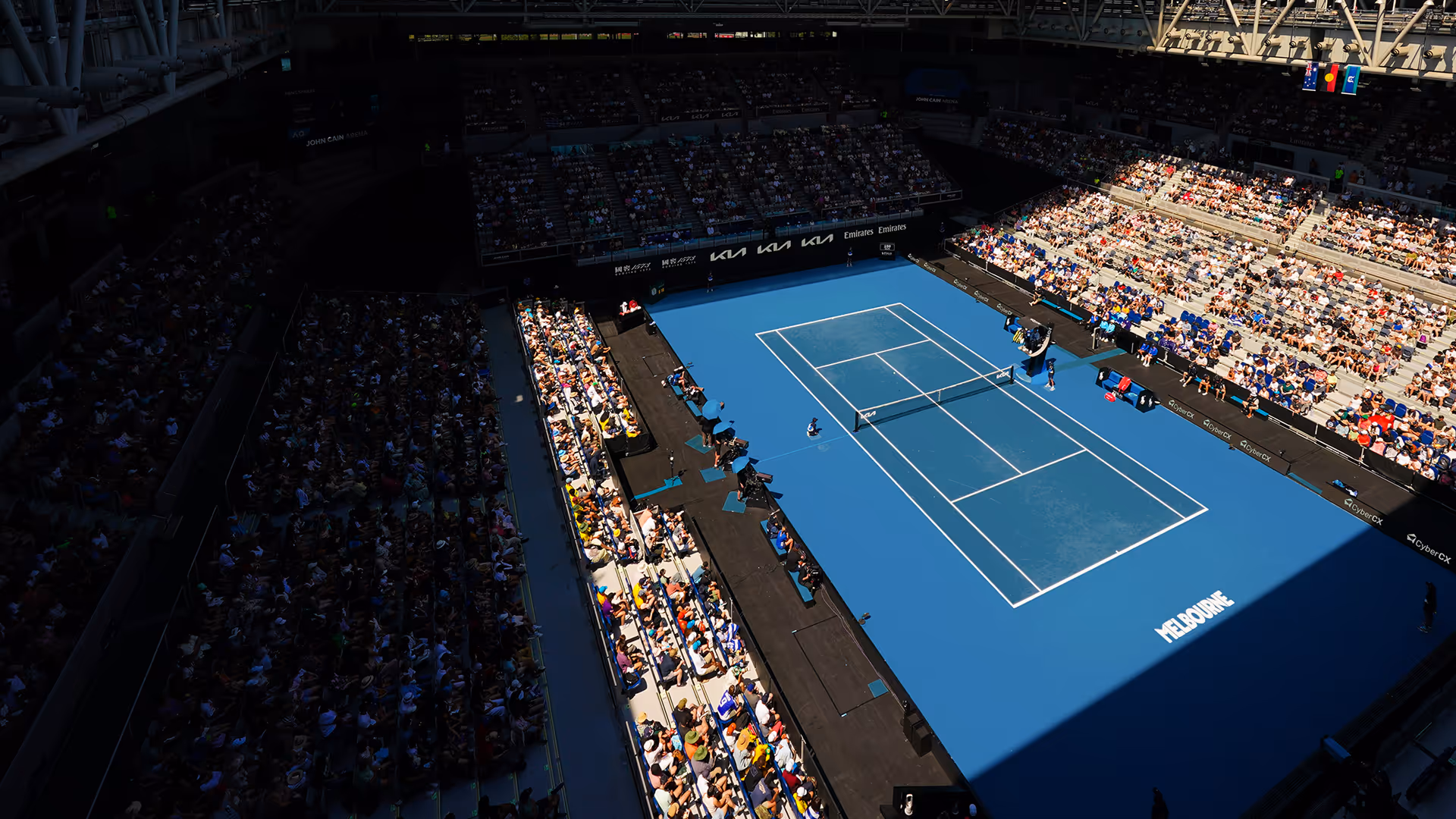 Aerial view of a tennis court with spectators in a stadium, featuring the word 'Melbourne' on the court.