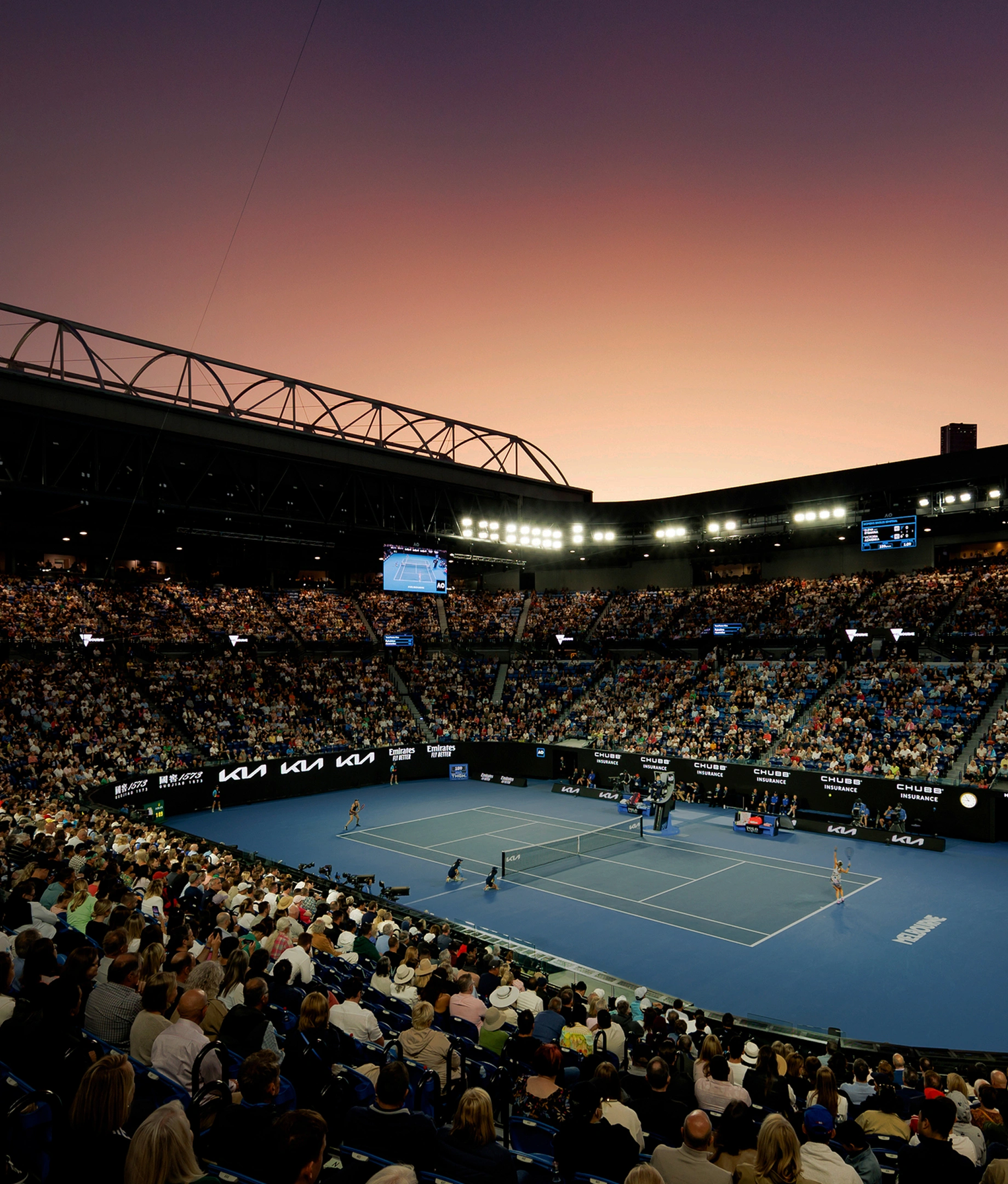 Wide view of a tennis match in a large stadium filled with spectators during sunset.
