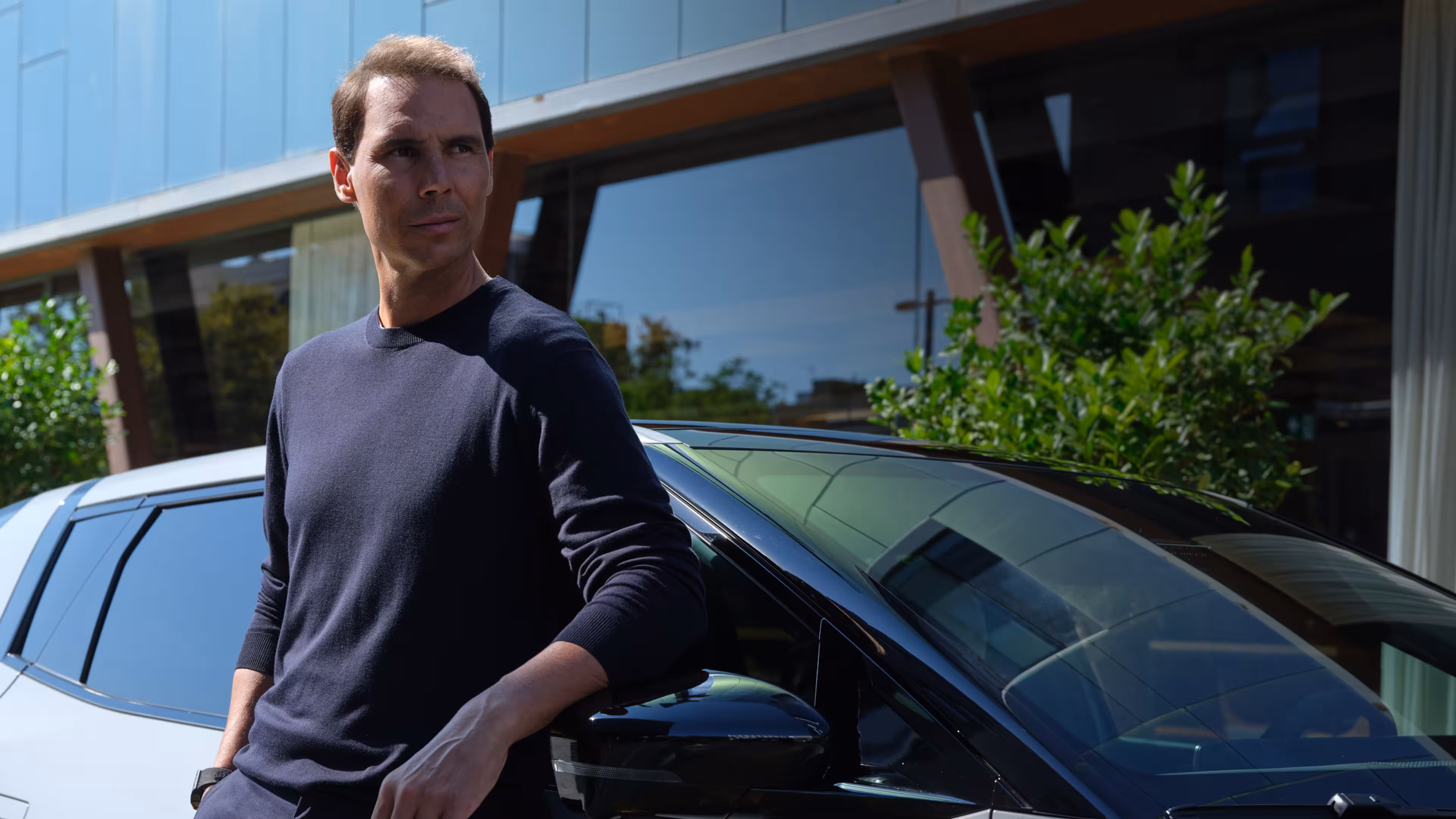 Man in a dark sweater leaning on a black car with a modern building and greenery in the background.