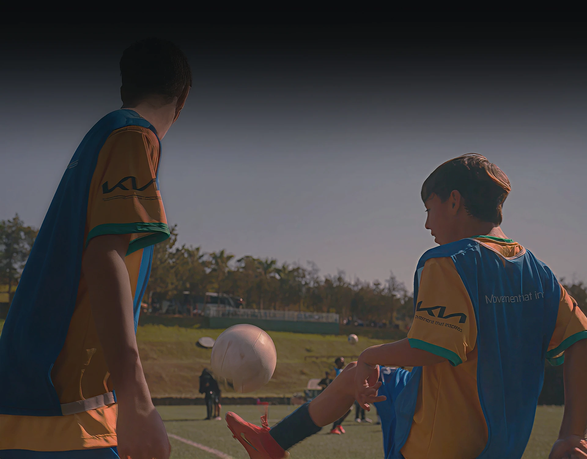 Two boys in sportswear on a field play soccer, one kicking a ball in mid-air during daytime.