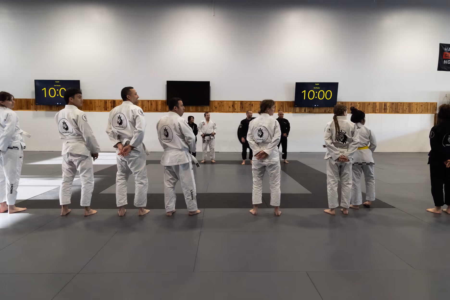 Group of Brazilian Jiu-Jitsu practitioners in white gis standing barefoot on gray mats in a training gym.