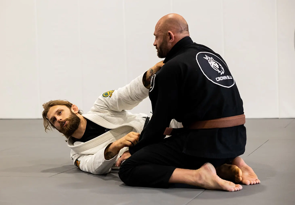 Two men practicing Brazilian Jiu-Jitsu on a mat, one in a white gi and the other in a black gi with a brown belt.