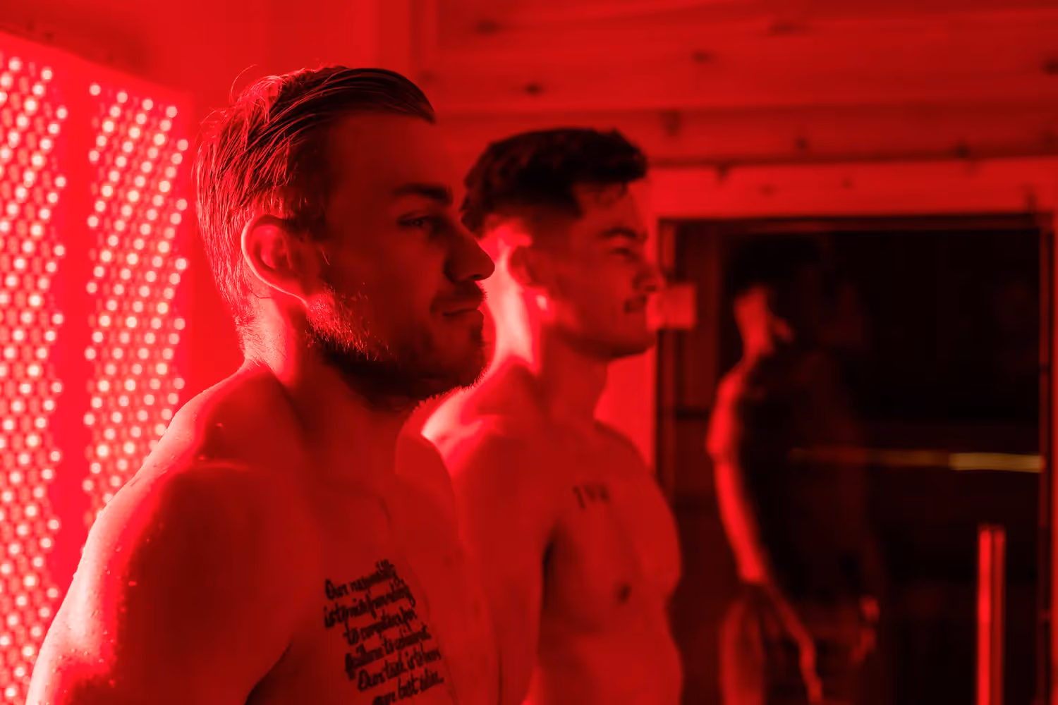 Two shirtless men standing side by side under red lighting, with one showing a chest tattoo.