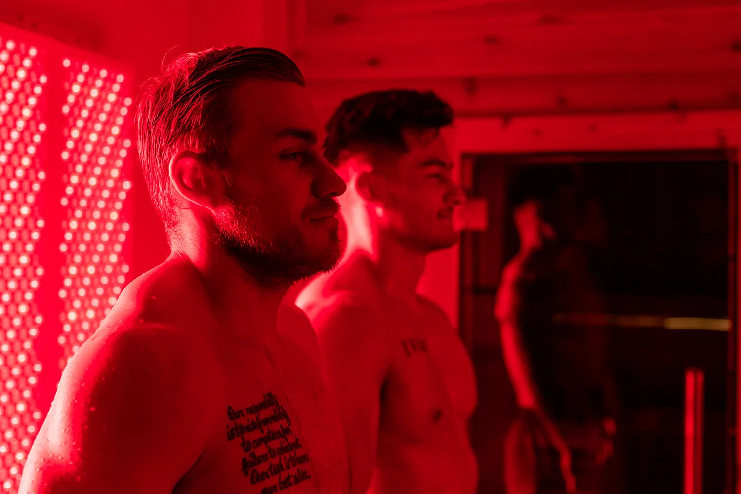 Two shirtless men standing side by side under red lighting, with one showing a chest tattoo.